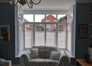 A tall window in a modern Victorian home. The shutters are a café style which exposes the glass above the shutters. The ceiling is white and the walls are blue. In front of the window is a large single seat sofa with a light blue throw over it. The sofa also has 3 scatter cushions.