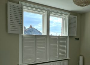 A Tier-on-Tier shutter. The top panels are open revealing the window glass and a blue sky. The louvers on the bottom section of the shutter are closed thus reducing the light. The walls are light clay in colour, and there is a radiator under the window.