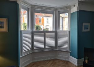 A tall window in a modern Victorian home. The shutters are a café style which exposes the glass above the shutters. The ceiling is white and the walls are blue. There is a wooden floor and 2 small pictures either side of the window.