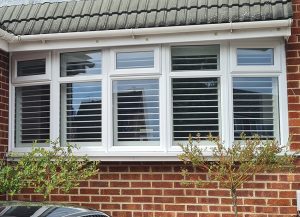 A Modern home with a bay window. Looking at the window from outside the house. The window frame is white and surrounded by red brick. There are 2 small green bushes growing up in front of the window