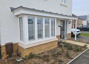 A large bay window on a modern house. The view is from the outside looking at the window. Under the window are red bricks and then above the house is rendered and painted white. The frame of the window is white and the shutters on the inside are visible from the outside. The garden is small and is not yet established so only mud and wood chippings are visible.